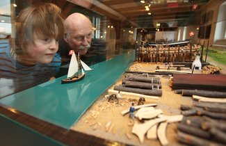 Ein Vater schaut sich gemeinsam mit seinem Sohn eine Vitrine im Flensburger Schifffahrtsmuseum an. In dieser ist ein Modell eines Schiffbauplatzes zu sehen. Im Vordergrund ist das Holzlager und ganz hinten werden Holzschiffe gebaut.