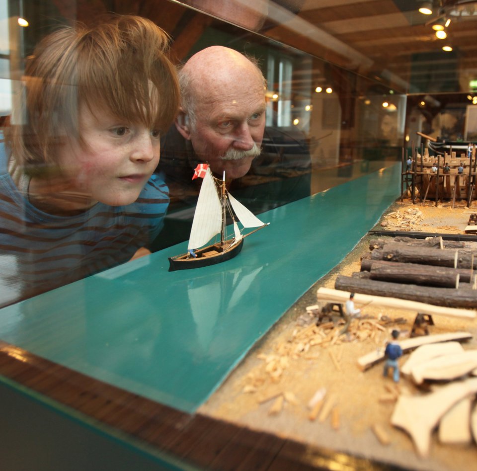 Ein Vater schaut sich gemeinsam mit seinem Sohn eine Vitrine im Flensburger Schifffahrtsmuseum an. In dieser ist ein Modell eines Schiffbauplatzes zu sehen. Im Vordergrund ist das Holzlager und ganz hinten werden Holzschiffe gebaut.