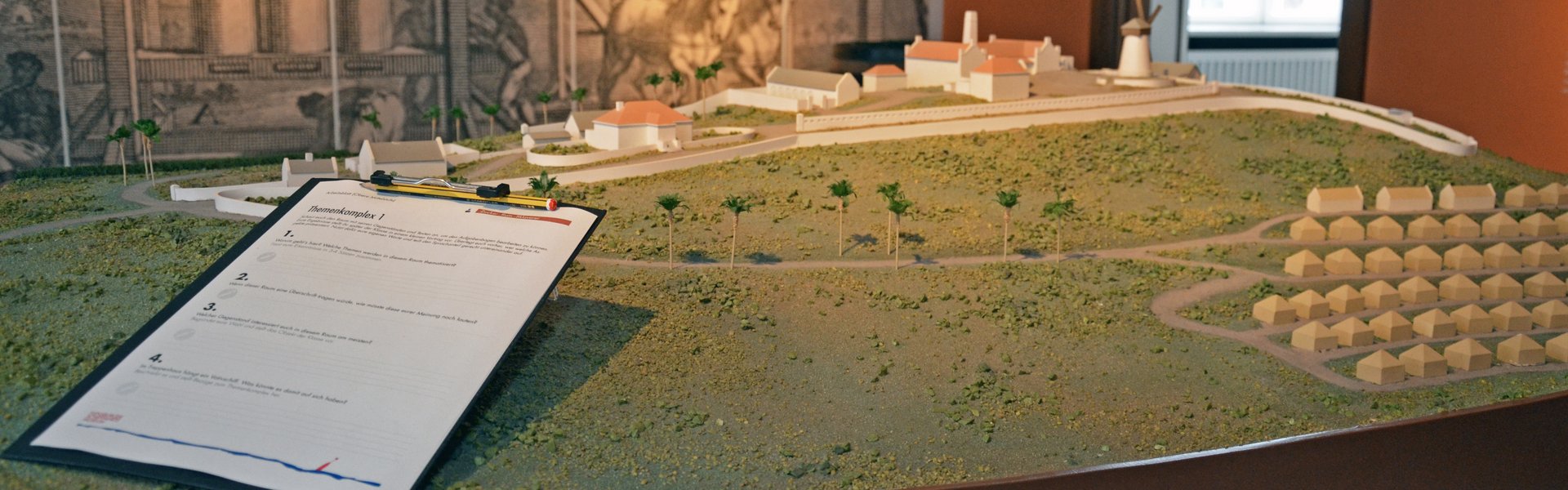 Farbige Fotografie, die einen Blick in die Ausstellung zeigt. In der Mitte des Raumes steht ein Modell. Es zeigt eine Zuckerrohr-Plantage mit dem Feld und vielen kleinen und ein paar großen Gebäuden. Auf dem Modell ist ein Klemmbrett abgelegt. Auf dem Klemmbrett ist ein Papier mit Fragen sowie ein Bleistift fixiert. Im Hintergrund der Fotografie ist eine Wandtapete abgebildet. Sie zeigt eine von Tieren betriebene Zuckermühle.