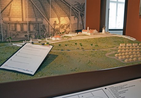 Farbige Fotografie, die einen Blick in die Ausstellung zeigt. In der Mitte des Raumes steht ein Modell. Es zeigt eine Zuckerrohr-Plantage mit dem Feld und vielen kleinen und ein paar großen Gebäuden. Auf dem Modell ist ein Klemmbrett abgelegt. Auf dem Klemmbrett ist ein Papier mit Fragen sowie ein Bleistift fixiert. Im Hintergrund der Fotografie ist eine Wandtapete abgebildet. Sie zeigt eine von Tieren betriebene Zuckermühle.