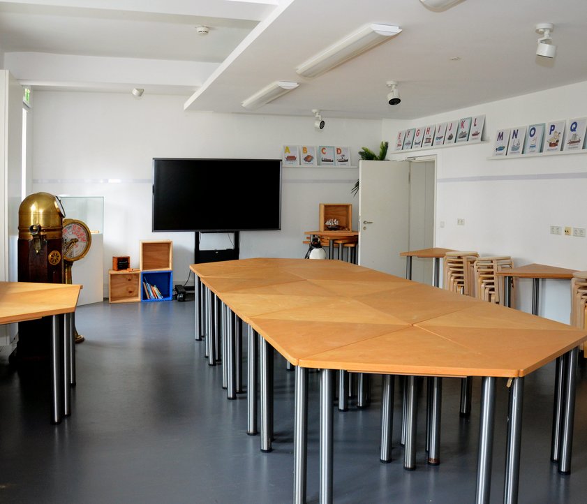 Blick in den Museumspädagogischen Raum des Schifffahrtsmuseums. Es ein schmaler, langer Raum. In der Mitte stehen Dreieckstische zusammengestellt zu einer langen Tafel. An den Seiten stehen weitere Tische und Hocker. Im Hintergrund steht ein Smartboard.