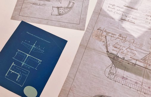 Auf einem weißen Tisch liegen verschiedene Bauzeichnungen für Schiffe. Eins der Papiere ist blau und die Zeichnung weiß. Beide anderen Papiere sind weiß.