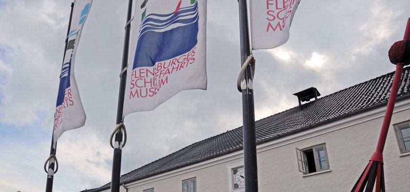 Drei Fahnenmasten mit Flaggen, die das Logo des Flensburger Schifffahrtsmuseum zeigen, stehen vor dem Museum. Das Logo ist eine grafische Darstellung des Gebäudes mit seinen zwei Toren und dem roten Seezeichen davor. Auch sind auf dem Logo Wellen zu sehen und der Schriftzug „Flensburger Schifffahrtsmuseum“.