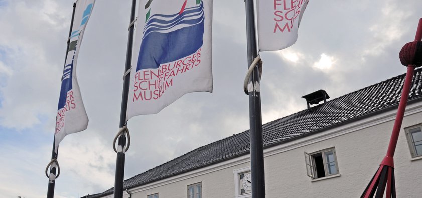 Foran museet står tre flagstænger med flag, der bærer Søfartsmuseets logo. Logoet er en grafisk gengivelse af bygningen med dens to porte og det røde søtegn foran. Logoet viser også bølger og teksten »Flensburger Schifffahrtsmuseum«.