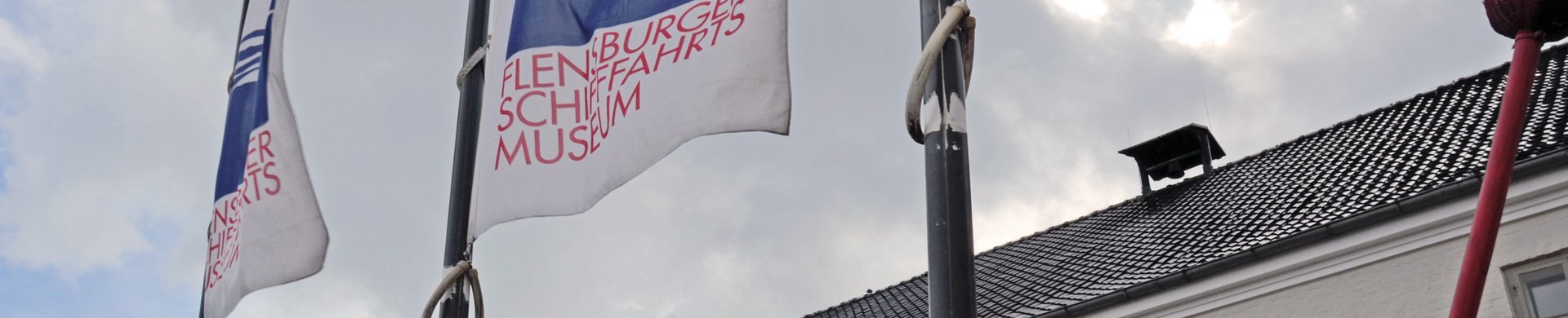 Drei Fahnenmasten mit Flaggen, die das Logo des Flensburger Schifffahrtsmuseum zeigen, stehen vor dem Museum. Das Logo ist eine grafische Darstellung des Gebäudes mit seinen zwei Toren und dem roten Seezeichen davor. Auch sind auf dem Logo Wellen zu sehen und der Schriftzug „Flensburger Schifffahrtsmuseum“.