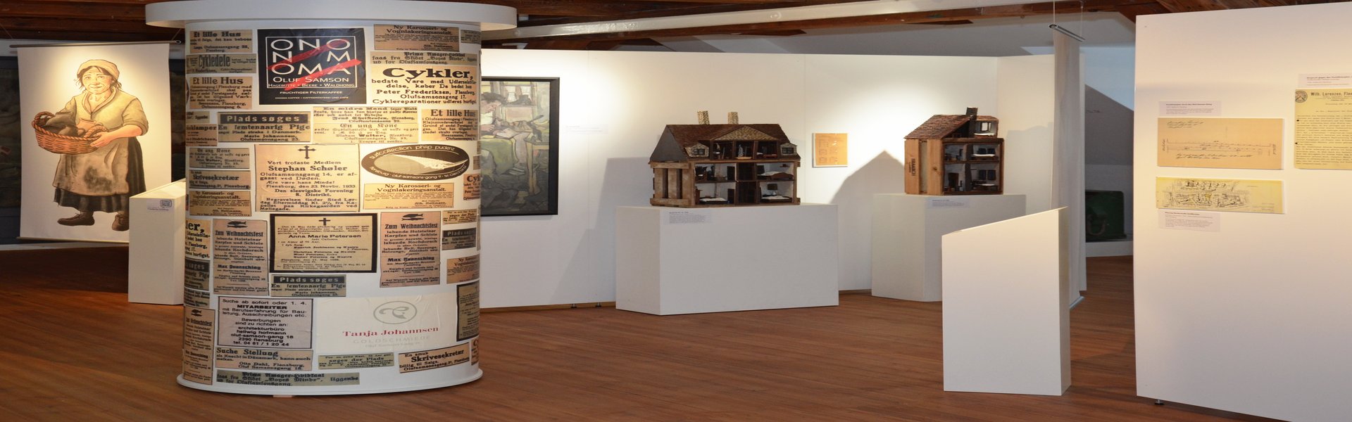 In der Mitte der Ausstellung IDYLLE UND KONFLIKT steht eine Litfaßsäule, die mit verschiedenen alten Zeitungsanzeigen beklebt ist. Alle handeln von dem Oluf-Samson-Gang. Dahinter stehen Modellhäuser zweier Häuser aus dem Gang. Sie stehen vor einer Stellwand. An dieser hängt ein Gemälde, das einen Schuster zeigt. Links im Hintergrund ist ein großes Banner zu sehen. Es zeigt eine mittelalte Frau, die in der Hand einen Korb mit Töpferware trägt. Ihre Kleidung ist schlicht und ein wenig schmutzig.