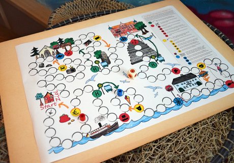 Ein Brettspiel liegt auf einem Fischernetz, das wiederum über einem Fass hängt. Auf dem Spielbrett sind zwei Figuren und ein Würfel platziert. Das Spielfeld besteht aus mehreren kreisrunden Feldern. Zudem sind verschiedene Zeichnungen rund um das Spielfeld gemalt.