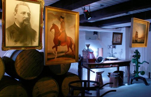 Im Hintergrund sind Rumfässer gestapelt. Vor diesen hängen zwei Bilder. Eins zeigt einen Mann mit Bart und Brille, ein anderes einen Mann, der auf einem Pferd sitzt. Zwei weitere Bilder hängen ganz links. Eines von ihnen zeigt ein Segelschiff auf dem Meer und das andere ein Segelschiff in der Nähe von Land. Alle diese Bilder stehen in Zusammenhang mit der Flensburger Rumgeschichte. Außerdem sind verschiedene Gefäße und Maschinen, die für die Rumabfüllung verwendet worden sind, auf dem Foto zu sehen.