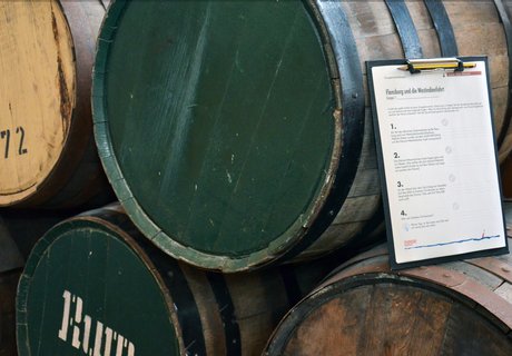 Auf der Farbfotografie sind Rumfässer zu sehen. Sie liegen und sind aufeinandergestapelt. Einige von ihnen haben einen grünen Boden, andere einen braunen. Auf einigen der Böden sind Zahlen gestempelt, auf einem das Wort Rum. Angelehnt an einem Rumfass steht ein Klemmbrett mit einem Aufgabebogen. Die Klammer hält nicht nur den Bogen, sondern auch einen Bleistift fest.