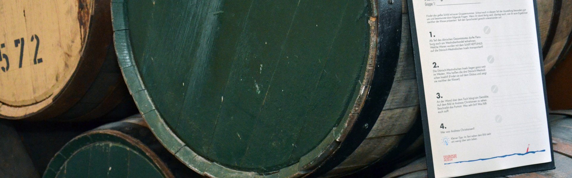 Auf der Farbfotografie sind Rumfässer zu sehen. Sie liegen und sind aufeinandergestapelt. Einige von ihnen haben einen grünen Boden, andere einen braunen. Auf einigen der Böden sind Zahlen gestempelt, auf einem das Wort Rum. Angelehnt an einem Rumfass steht ein Klemmbrett mit einem Aufgabebogen. Die Klammer hält nicht nur den Bogen, sondern auch einen Bleistift fest.