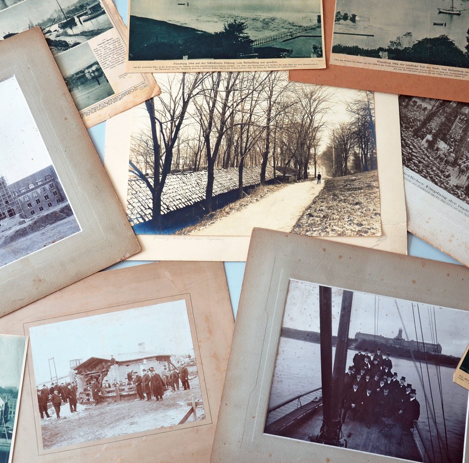 Verschiedene alte Fotos und Zeitungsartikeln mit Bildern liegen auf einem Tisch. Einige der Bilder haben ein Passepartout. Keines von ihnen ist jedoch gerahmt.