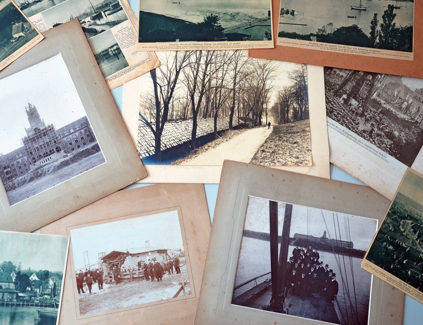 Verschiedene alte Fotos und Zeitungsartikeln mit Bildern liegen auf einem Tisch. Einige der Bilder haben ein Passepartout. Keines von ihnen ist jedoch gerahmt.