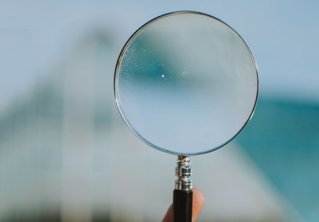 En lup holdes i hånden. Man kan kun se en enkelt finger af hånden. Baggrunden er helt sløret. Man kan kun ane en hvid bygning.