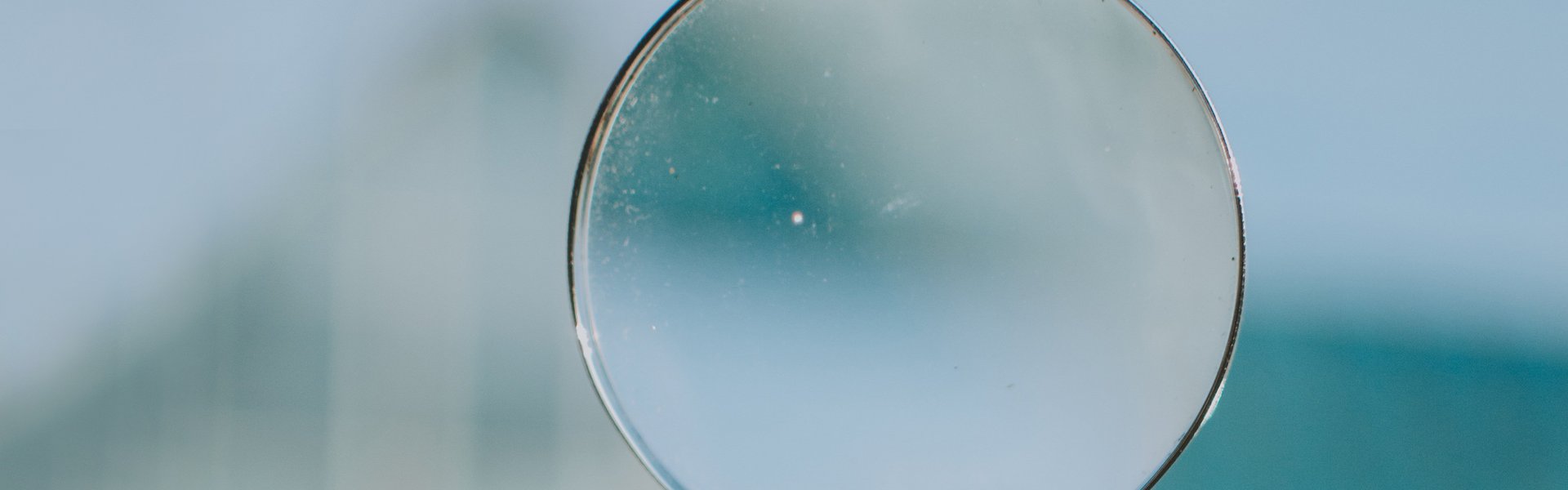 Eine Lupe wird in der Hand gehalten. Von der Hand sieht man nur einen einzigen Finger. Der Hintergrund ist total verschwommen. Man kann lediglich ein weißes Gebäude erahnen.