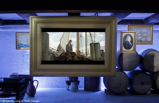 Auf einem großen Monitor, der von der Decke im Rum-Keller hängt, wird ein Film gezeigt. In der Szene sieht man einen jungen Mann, der auf der Reling eines alten Segelschiffes sitzt. Das Segelschiff ist an Dalben fest vertäut. Neben dem Monitor hängen drei Gemälde mit Goldrahmen. Sie zeigen Personen und Segelschiffe. Links und rechts von zentrierten Monitor stehen Fässer und andere Gefäße zur Rumaufbewahrung.