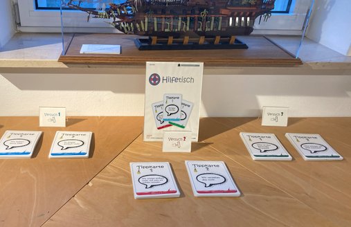 Eine bunte Fotografie. Darauf zu sehen ist der Ausschnitt eines Tisches. In der Mitte des Tisches befindet sich ein Aufsteller mit einem Schild, das die Aufschrift „Hilfetisch“ hat. Unter der Schrift sieht man die Abbildung von verschiedenen „Tippkarten“. Vor dem Schild befindet sich ein kleiner Aufsteller mit dem Text „Versuch 2“. Davor liegen zwei Stapel mit Karten. Links und rechts von dem Aufsteller in der Mitte befinden sich ebenfalls jeweils ein kleiner Aufsteller und zwei Stapel Karten davon. Im Hintergrund sieht man den Teil einer Fensterbank. 
