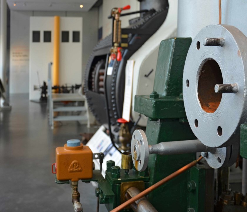 Blick durch die Technikhalle. Rechts und links an den Wänden stehen verschiedene Maschinen. Auch im Vordergrund ist eine große Maschine zu erkennen. Im Hintergrund ist die Brücke eines Schiffes angedeutet.