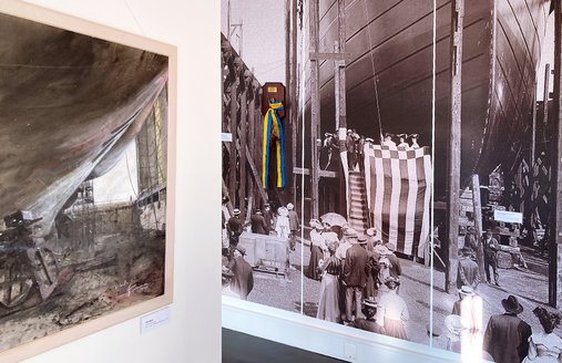 På en skillevæg hænger et moderne maleri af et værft. Det viser et skib under bygning. På væggen til højre hænger et fototapet. Det viser en historisk skibsdåb.