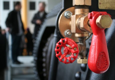 Im Vordergrund ist der Dampfkessel eines Schiffes zu sehen. Im Hintergrund stehen zwei Personen und unterhalten sich miteinander. 