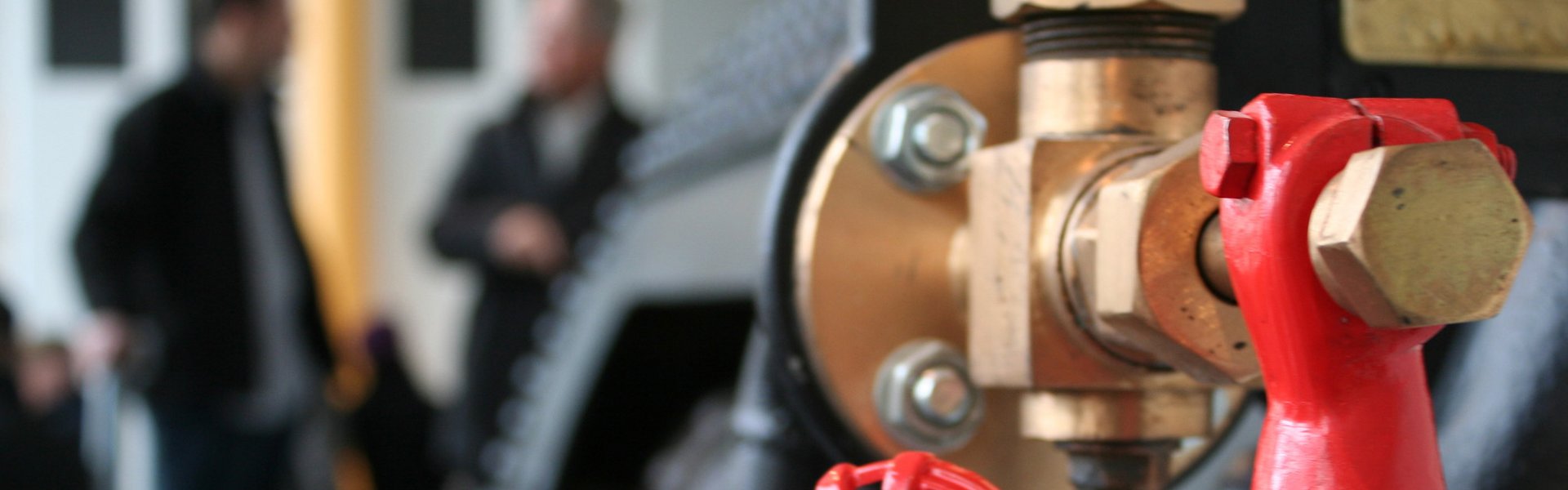 Im Vordergrund ist der Dampfkessel eines Schiffes zu sehen. Im Hintergrund stehen zwei Personen und unterhalten sich miteinander. 