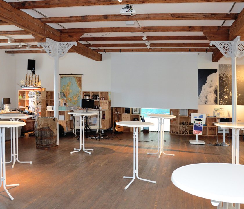 Blick ins Foyer des Flensburger Schifffahrtsmuseums. Stehtische sind aufgestellt und im Hintergrund eine Leinwand von der Decke heruntergelassen. Daneben steht ein Stehpult.