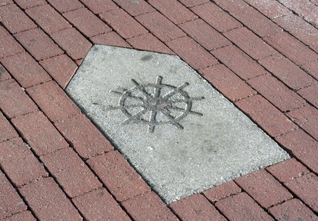 Der Wegweiser für den KAPITÄNSWEG ist im Pflaster überall in der Stadt eingelassen.  Der Wegweiser ist selbst ein Stein in Pfeilform mit einem Steuerrad, das in den Stein reingeritzt worden ist. Dieser Wegweiser ist von roten Pflastersteinen umgeben.