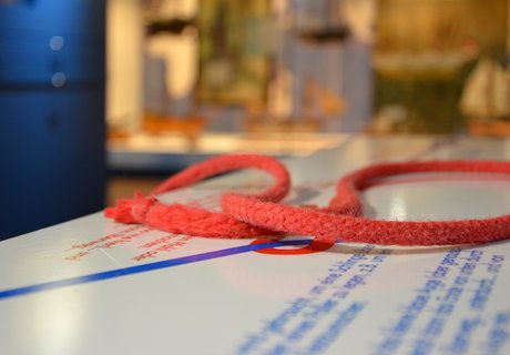 Auf einer Knotentafel liegt ein roter Tampen. Dieser ist zu einer Schlaufe gelegt. Im Hintergrund ganz verschwommen ist eine große Vitrine mit Schiffsmodellen zu sehen und eine senkrechte blaue Stele.