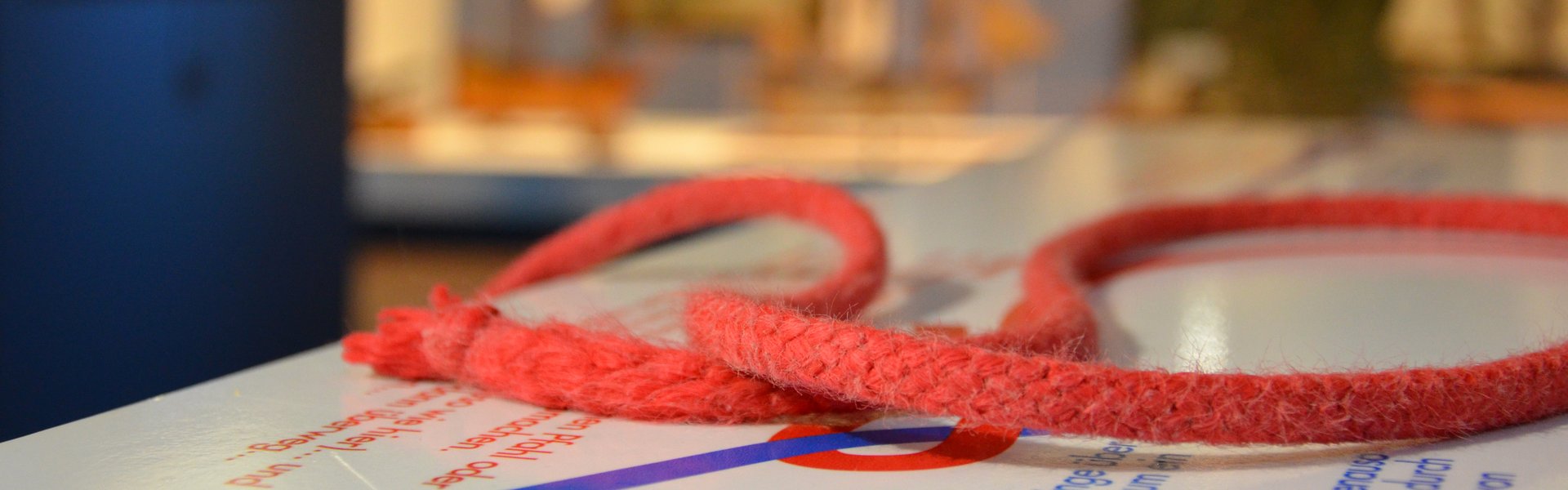 Auf einer Knotentafel liegt ein roter Tampen. Dieser ist zu einer Schlaufe gelegt. Im Hintergrund ganz verschwommen ist eine große Vitrine mit Schiffsmodellen zu sehen und eine senkrechte blaue Stele.