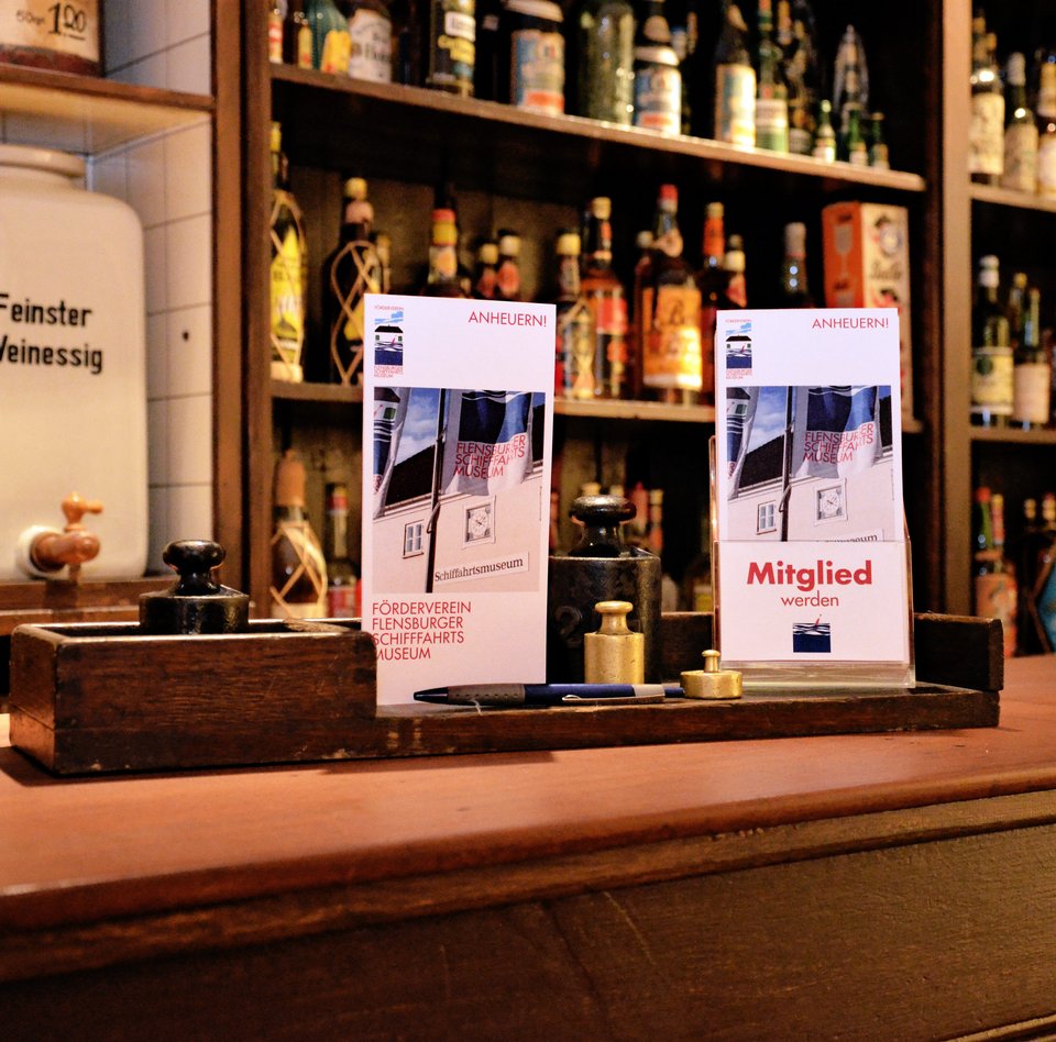 Billedet viser C.C. Petersens kolonivarerbutik. Den findes i udstillingen „Rom.Sukker.Slaveri.“ i Søfartsmuseets bagbygning. På disken ligger tilmeldingsblanketter til medlemskab af støtteforeningen.