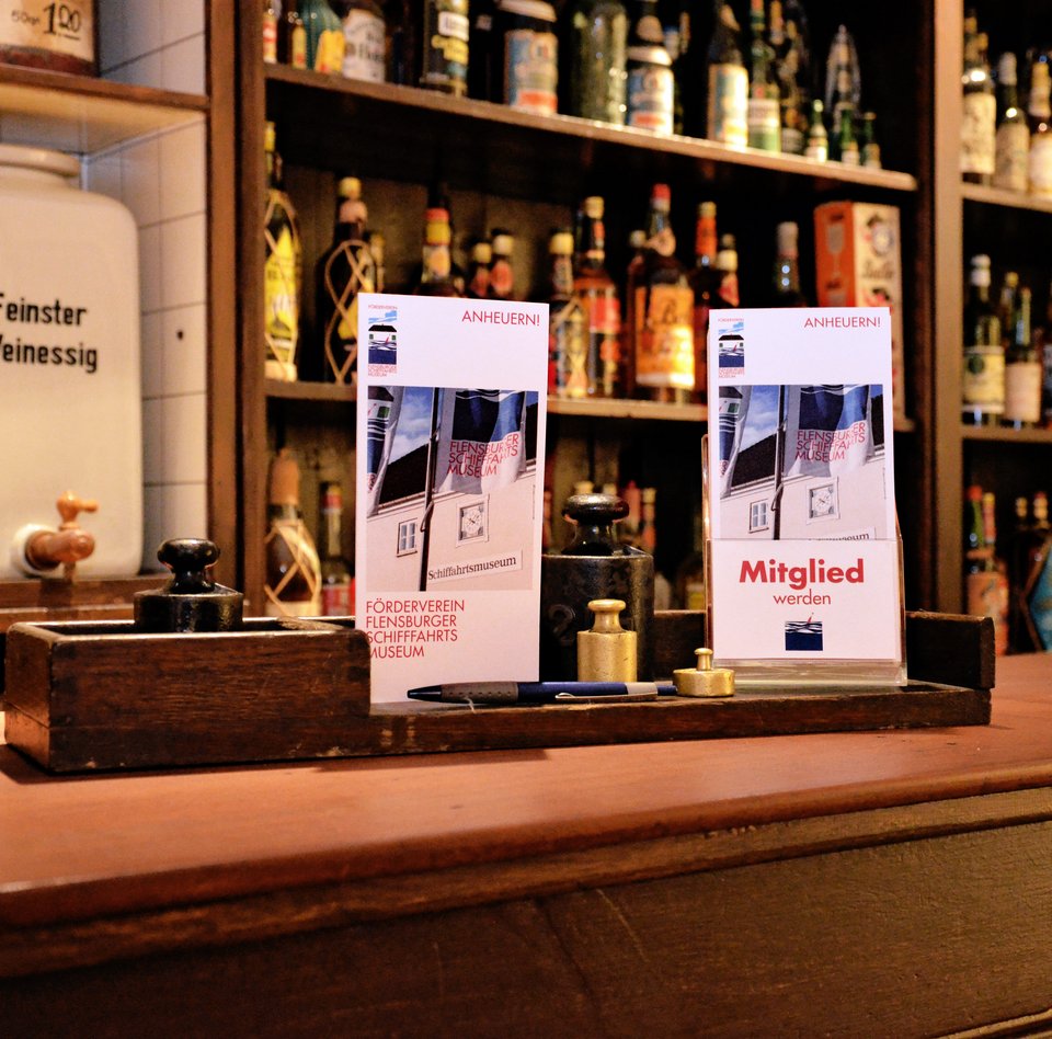 Das Bild zeigt den Kolonialwarenladen von C.C. Petersen. Dieser befindet sich in der Ausstellung "Rum.Zucker.Versklavung." im Hintergebäude des Schifffahrtmuseums. Auf dem Tresen sind Anmeldeformulare für die Mitgliedschaft im Förderverein.