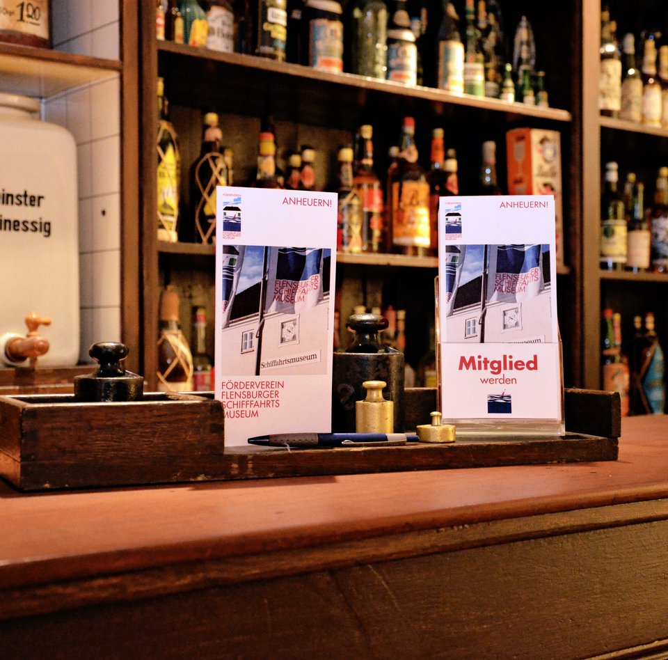 Das Bild zeigt den Kolonialwarenladen von C.C. Petersen. Dieser befindet sich in der Ausstellung "Rum.Zucker.Versklavung." im Hintergebäude des Schifffahrtmuseums. Auf dem Tresen sind Anmeldeformulare für die Mitgliedschaft im Förderverein.