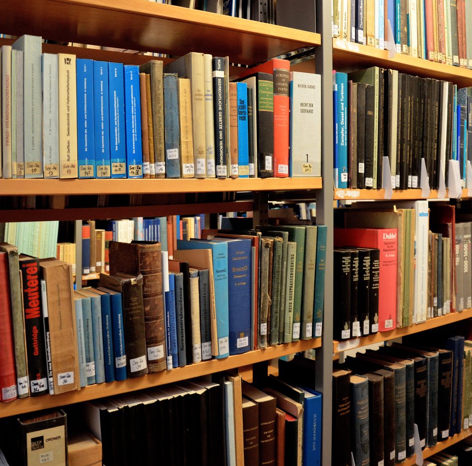 Regale aus Holz und Metall stehen dicht an dicht. In den Regalen stehen verschiedene Bücher in Reih und Glied – hierunter Fachbücher über Seefahrt aber auch Jahresmagazine von der Flensburger Werft. Alle Bücher haben einen kleinen weißen Aufkleber auf dem Rücken. Auf diesem steht eine Signatur. Über diese Signatur kann das Buch im Online-Verzeichnis des Museums vom Museumsteam gefunden werden.