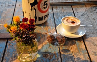 Auf einer alten Kiste steht eine Blumenvase, daneben eine Lampe. Auch eine Tasse Cappuccino und zwei Rumkugeln in einer Tüte sind auf der Kiste, die im Café des Schifffahrtsmuseums als eine Art Tresen dient. 