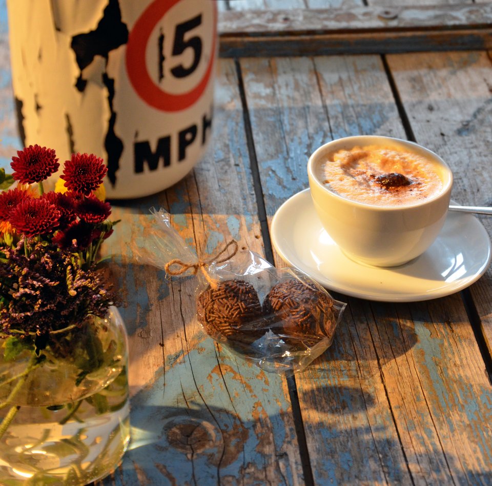 På en gammel kasse står en vase med blomster og ved siden af en lampe. Der står også en kop cappuccino og to romkugler i en pose på kassen, der fungerer som en slags disk i caféen på Søfartsmuseet.