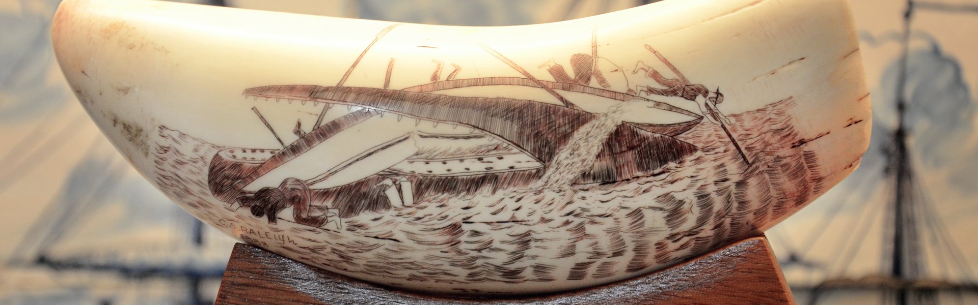 Im Vordergrund der Fotografie ist ein Walzahn zu sehen. Dieser ist auf einem hölzernen Sockel drapiert. Auf dem Zahn ist eine Walfangsszene gezeichnet. Sie zeigt, wie ein Wal ein Schiff im Maul hat und dieses durchbeißt. Ein Besatzungsmitglied fällt ins Wasser. Im Hintergrund der Fotografie ist ein Kachelbild zu sehen. Es ist in den Farben weiß und blau gehalten.