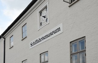 An der Fassade des ehemaligen Zollpackhauses in Flensburg befindet sich eine große Uhr. Unter dieser hängt ein Schild, auf dem „Schiffahrtsmuseum“ steht. 
