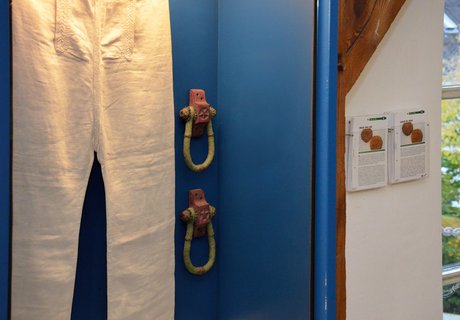 Auf dem farbigen Foto ist eine Seemannshose in einer blauen hängenden Vitrine zu sehen. Daneben – ebenfalls an der Wand – hängen zwei durchsichtige Kartenbehälter. In diesen sind Sammelkarten einsortiert. Diese bestehen aus einem Bild und Text.