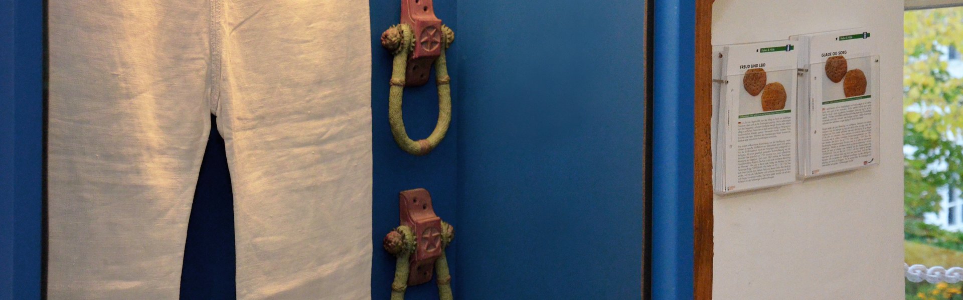 Auf dem farbigen Foto ist eine Seemannshose in einer blauen hängenden Vitrine zu sehen. Daneben – ebenfalls an der Wand – hängen zwei durchsichtige Kartenbehälter. In diesen sind Sammelkarten einsortiert. Diese bestehen aus einem Bild und Text.