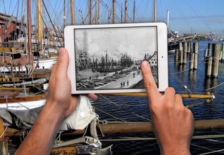  Im Vordergrund sind Hände zu sehen, die ein Tablet hochhalten und bedienen. Auf dem Tablet ist eine alte, gezeichnete Hafenansicht zu sehen. Im Hintergrund verschwommen ist der heutige Museumshafen zu sehen.
