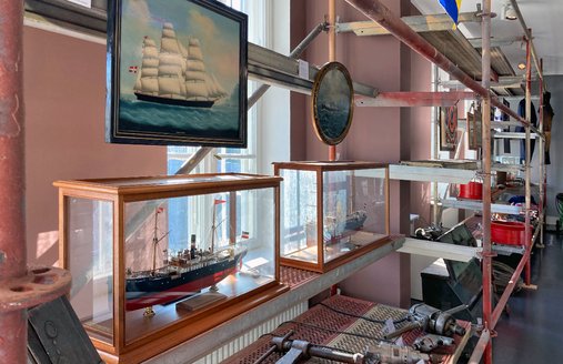 Auf dem Foto ist das lange Metallregal abgebildet. Es ist perspektiv aufgenommen, sodass eine diagonale Flucht im Bild entsteht. Gut zu sehen sind zwei Schiffsportraits, die im Regal hänge. Es handelt sich bei den Schiffen um die ersten Schiffe, die die FSG gebaut hat. Es sind Holzschiffe, die Segel haben. Später baute die FSG auch Stahlschiffe. Zwei von ihnen stehen in Vitrinen auf dem mittleren Regalboden.