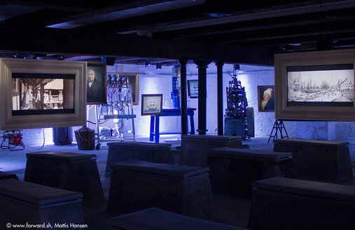 Im Vordergrund des Fotos sind Betonbänke zu sehen. Diese dienen als Sitzmöglichkeit während der Filmvorführung im Rum-Keller. Der Film wird auf Monitoren gezeigt. Auf diesem Foto kann man zwei der drei Monitore sehen, die von der Decke herunterhängen.