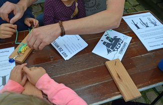 Kinder sitzen um einen Tisch. Auf diesem liegt eine Anleitung für die Herstellung eines Schlüsselanhängers aus Paracord. Dafür nutzen die Kinder eine Holzplatte, in die ganz oben in der Mitte ein Nagel eingeschlagen ist. Dort wird ein Schlüsselring befestigt und dann an diesen das Paracord geknotet.