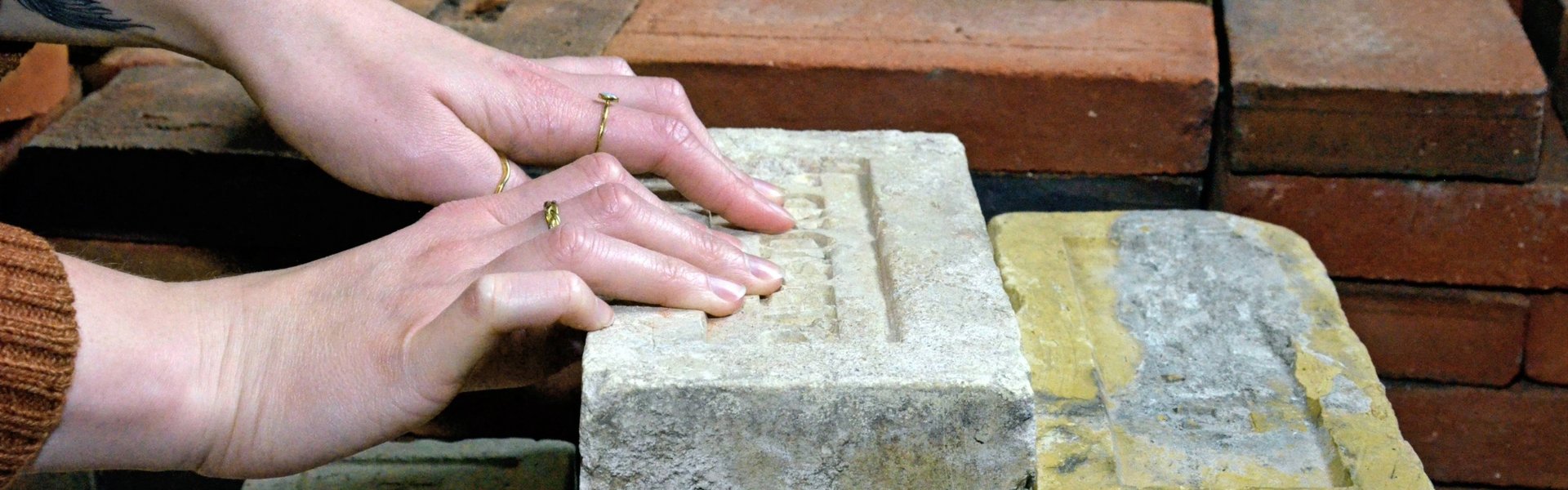 Auf dem Farbfoto sind Ziegelsteine zu sehen. Sie haben unterschiedliche Größen und Farben. Einige sind weiß, andere gelb und wieder andere rot. Die Ziegelsteine sind aufeinandergestapelt. Es gibt mehrere Stapel. Zwei Hände ertasten einen der größeren Steine. Auf dem Stein ist ein Wort eingemeißelt.