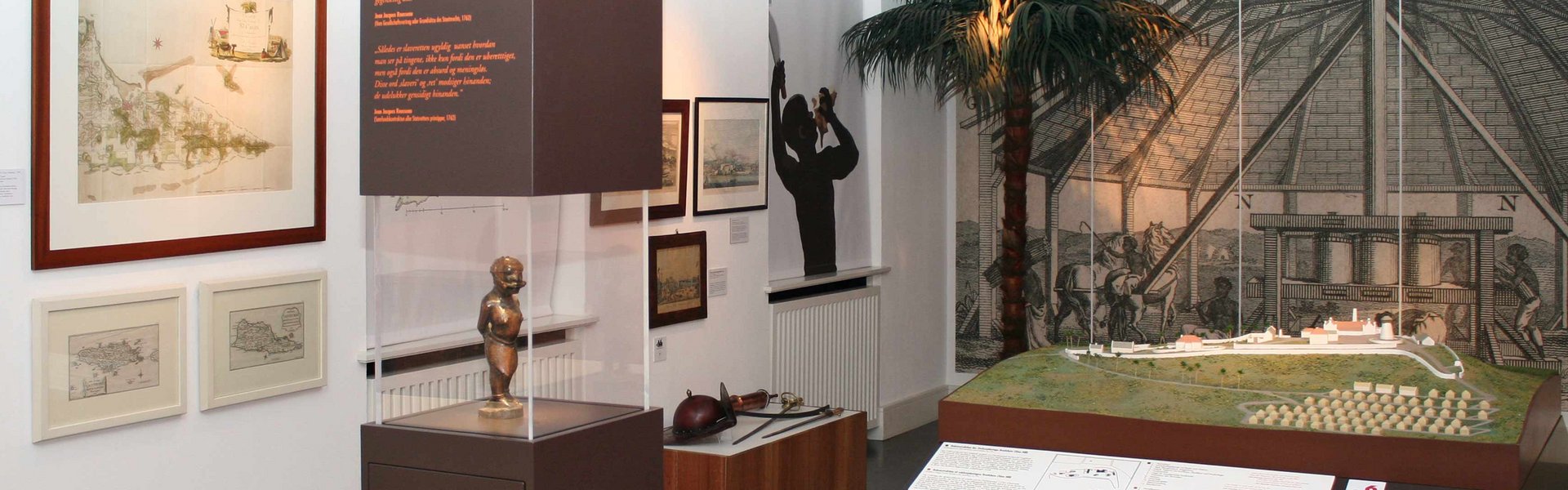 In der Mitte des Raumes steht eine braune Säule mit einer mittigen Glaseinfassung. Dort hinter ist eine Figur zu sehen, die einen versklavten Menschen aus Afrika darstellen soll. Dieser ist gefesselt. Dahinter steht ein großes Plantagenmodell mit kleinen Hütten und größeren Gebäuden für die Verarbeitung von Zuckerrohr. Im Hintergrund ist eine Wandtapete zu sehen. Auf dieser ist eine Zuckermühle abgebildet.