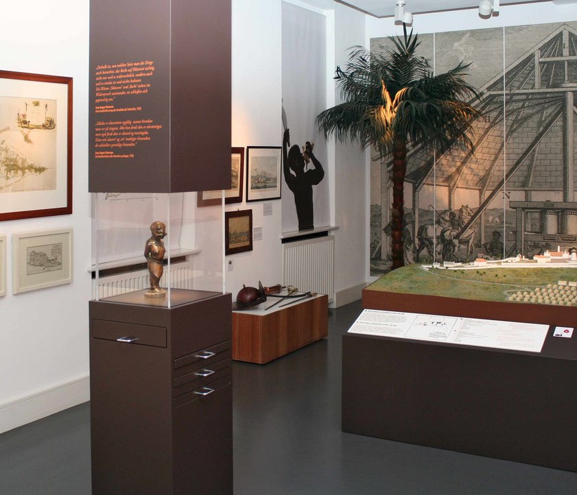 In der Mitte des Raumes steht eine braune Säule mit einer mittigen Glaseinfassung. Dort hinter ist eine Figur zu sehen, die einen versklavten Menschen aus Afrika darstellen soll. Dieser ist gefesselt. Dahinter steht ein großes Plantagenmodell mit kleinen Hütten und größeren Gebäuden für die Verarbeitung von Zuckerrohr. Im Hintergrund ist eine Wandtapete zu sehen. Auf dieser ist eine Zuckermühle abgebildet.