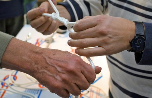 Über einer Knotentafel auf der die gängigsten Seemannsknoten abgebildet sind, wird mit Anleitung gerade ein Knoten gebunden. Es sind zwei jüngere und eine ältere Hand zu sehen.