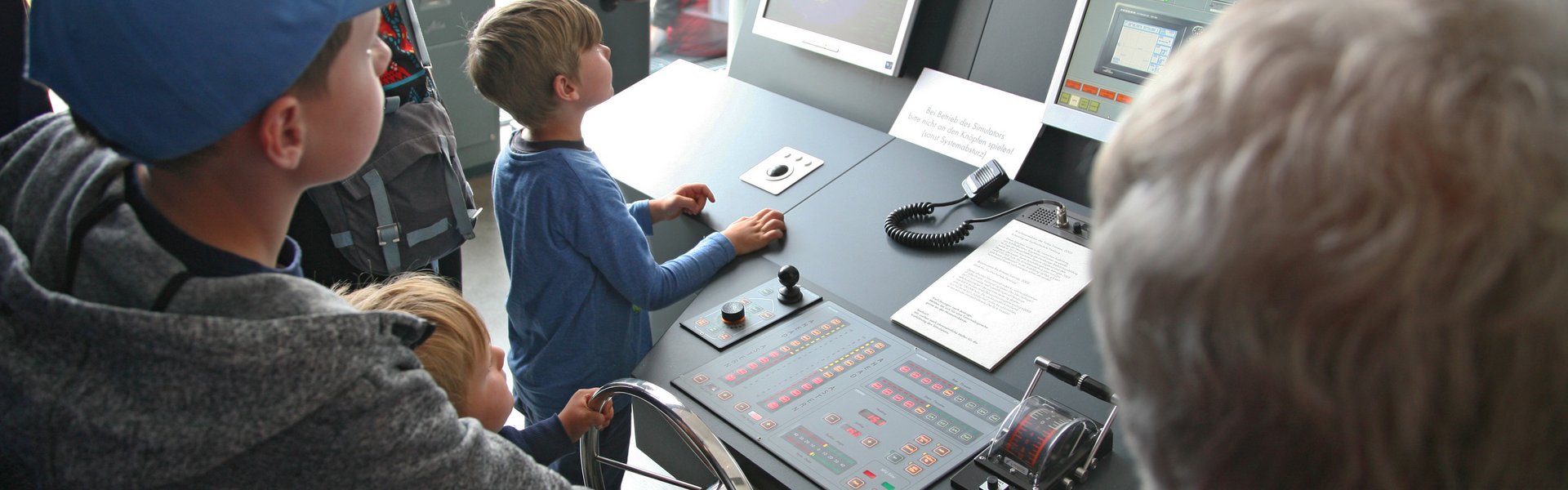 Auf dem farbigen Foto sind fünf Personen abgebildet: zwei Erwachsene und drei Kinder. Die Kinder stehen direkt am Schiffsführungssimulator. Zwei von ihnen halten das Ruder. Unterstützt werden sie dabei von einem weißhaarigen Mann.