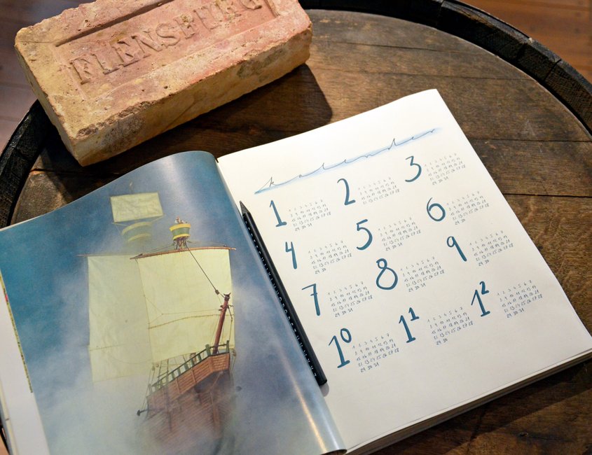Auf einem Fass liegt ein Terminkalender. Dieser ist aufgeschlagen und eine Jahresübersicht ist zu sehen. Daneben liegt ein Ziegelstein mit dem Aufdruck „Flensburg“.