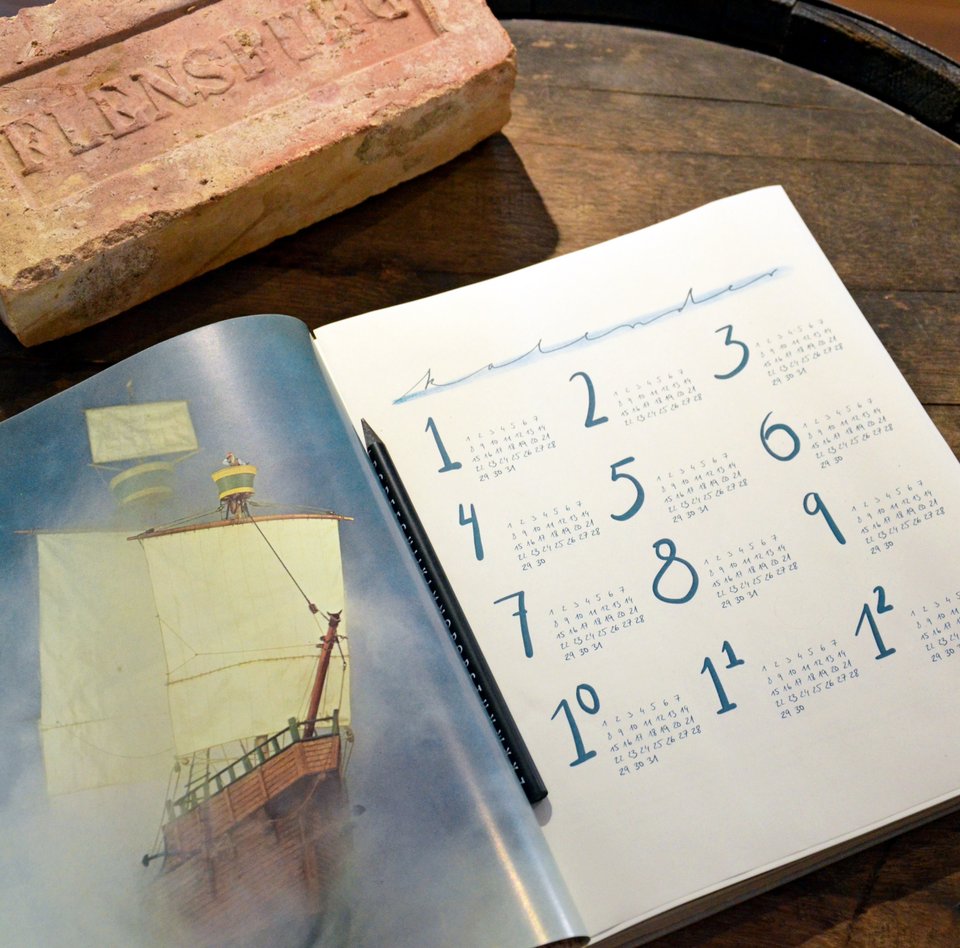 Auf einem Fass liegt ein Terminkalender. Dieser ist aufgeschlagen und eine Jahresübersicht ist zu sehen. Daneben liegt ein Ziegelstein mit dem Aufdruck „Flensburg“.