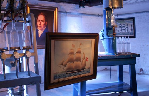 Das Schiffsportrait im Rum-Keller des Flensburger Schifffahrtsmuseums ist eingerahmt von einer Maschine zum Abfüllen von Rum und einer die zum Verkorken gedacht ist.